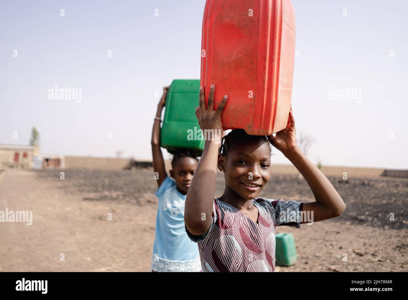 Close-up of two African girls carrying plastic water canisters on their heads and walking towards the village well on a dirt road Stock Photo