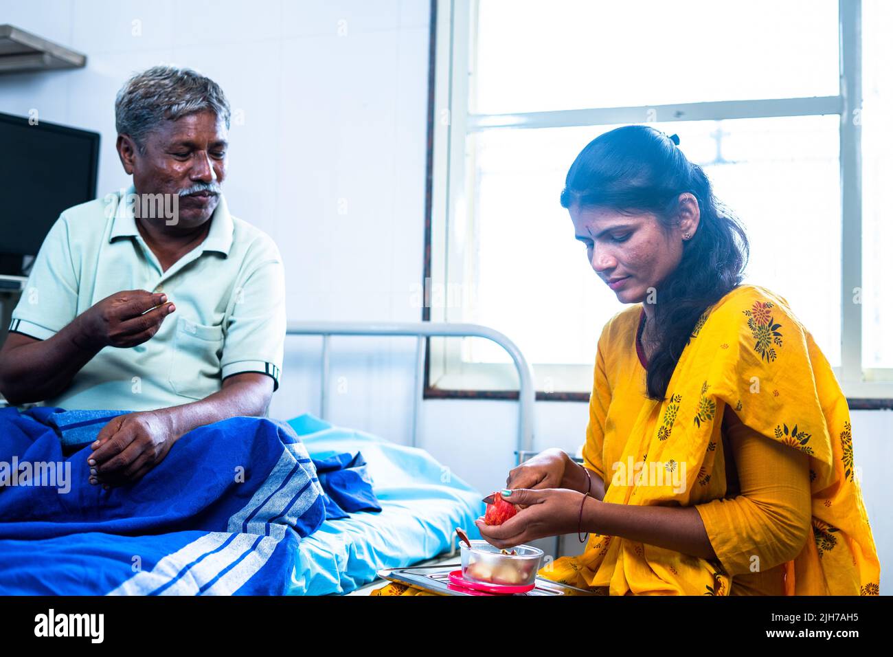 Happy smiling daughter giving fruits to recovered sick father while on bed at hospital - concept of health care, bonding and family support. Stock Photo