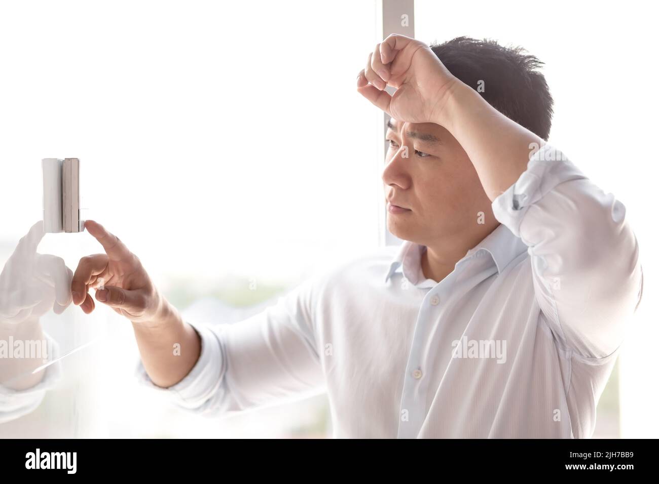 Close-up photo. A young handsome Asian man in a white shirt turns the air conditioner control button on, which hangs on the wall. Standing, leaning against the wall, very hot Stock Photo