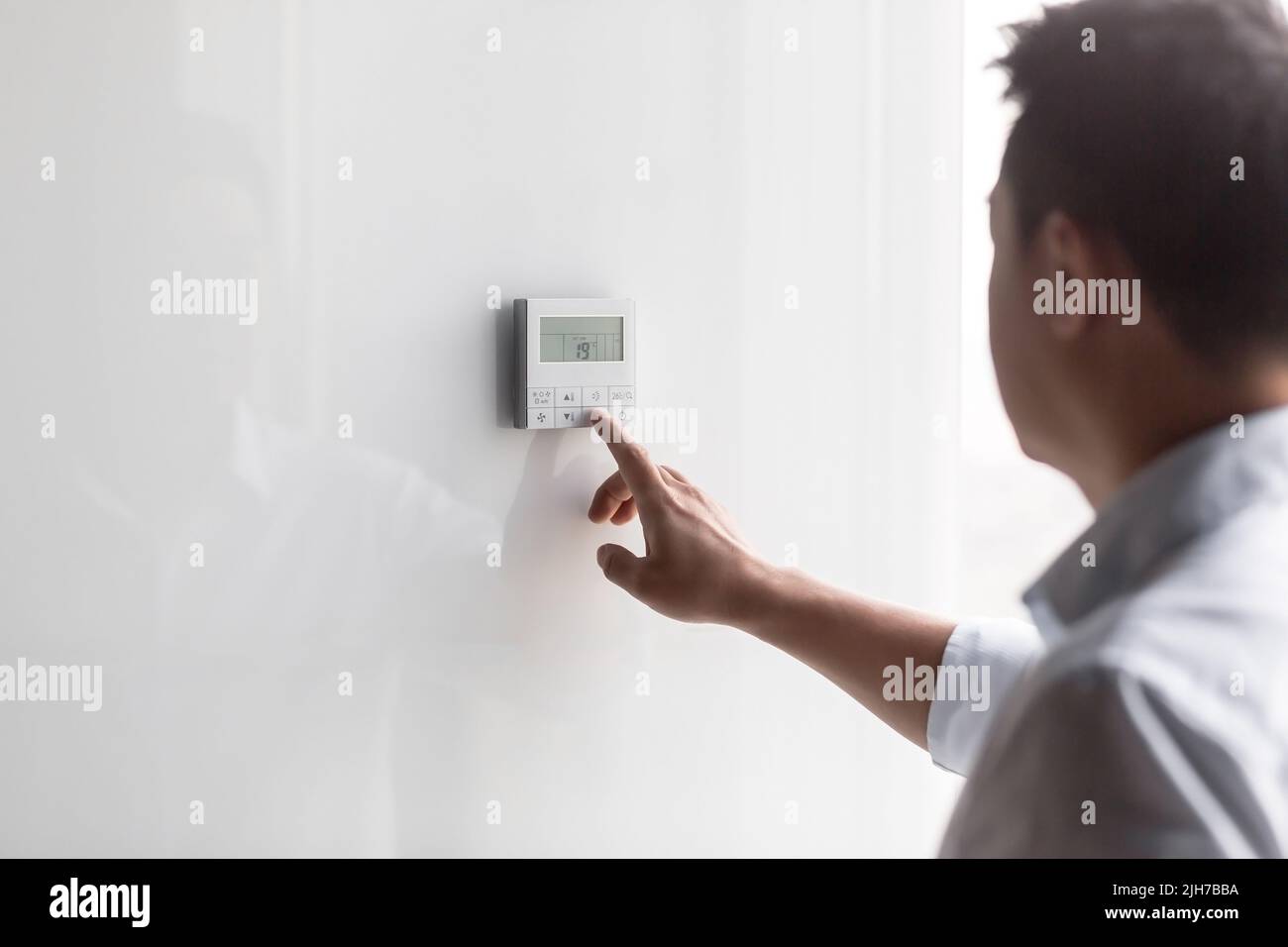 Close-up photo. The hand of a young man in a white shirt turns on the control buttons of the air conditioner hanging on the wall. Standing on the right Stock Photo