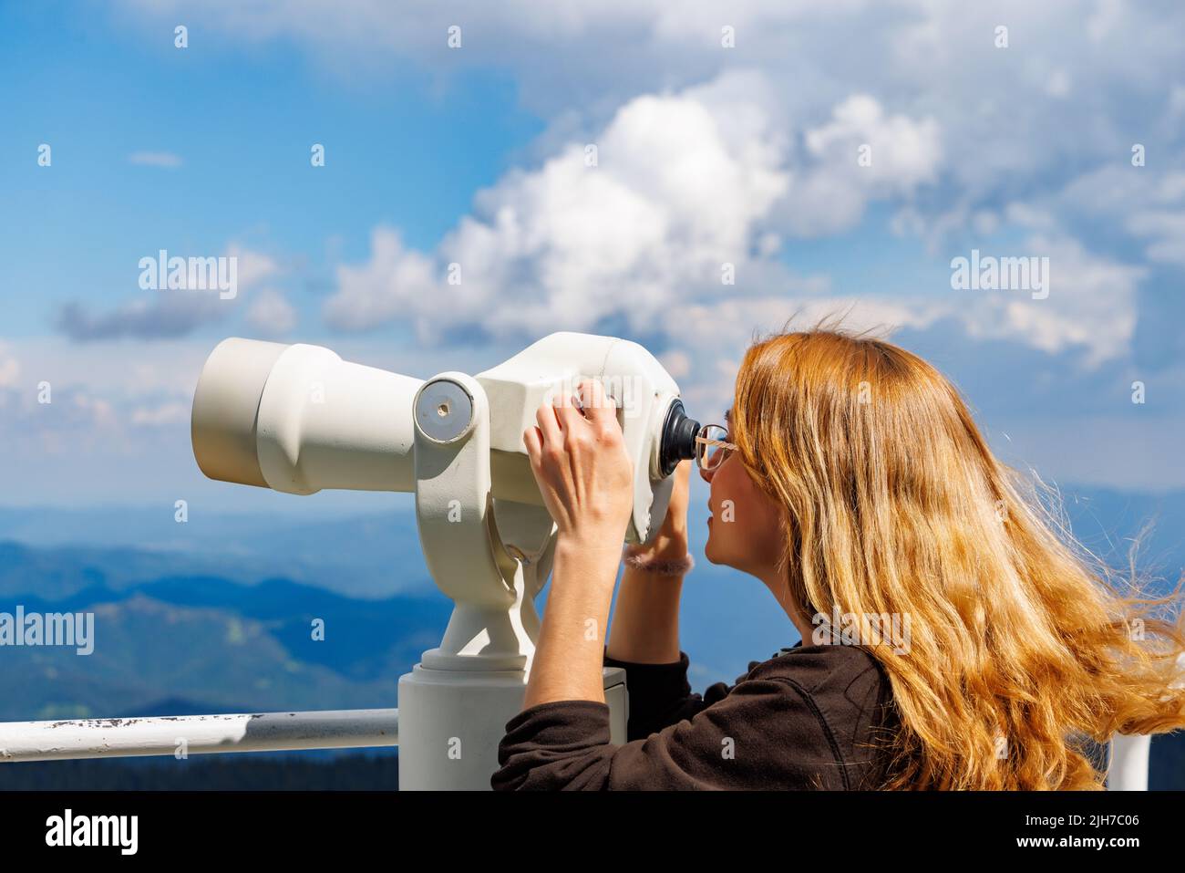 Curious girl tourist looks at mountainous wooded landscapes of mountain valley of Rhodope Mountains and blue cloudy sky through modern metal telescope Stock Photo