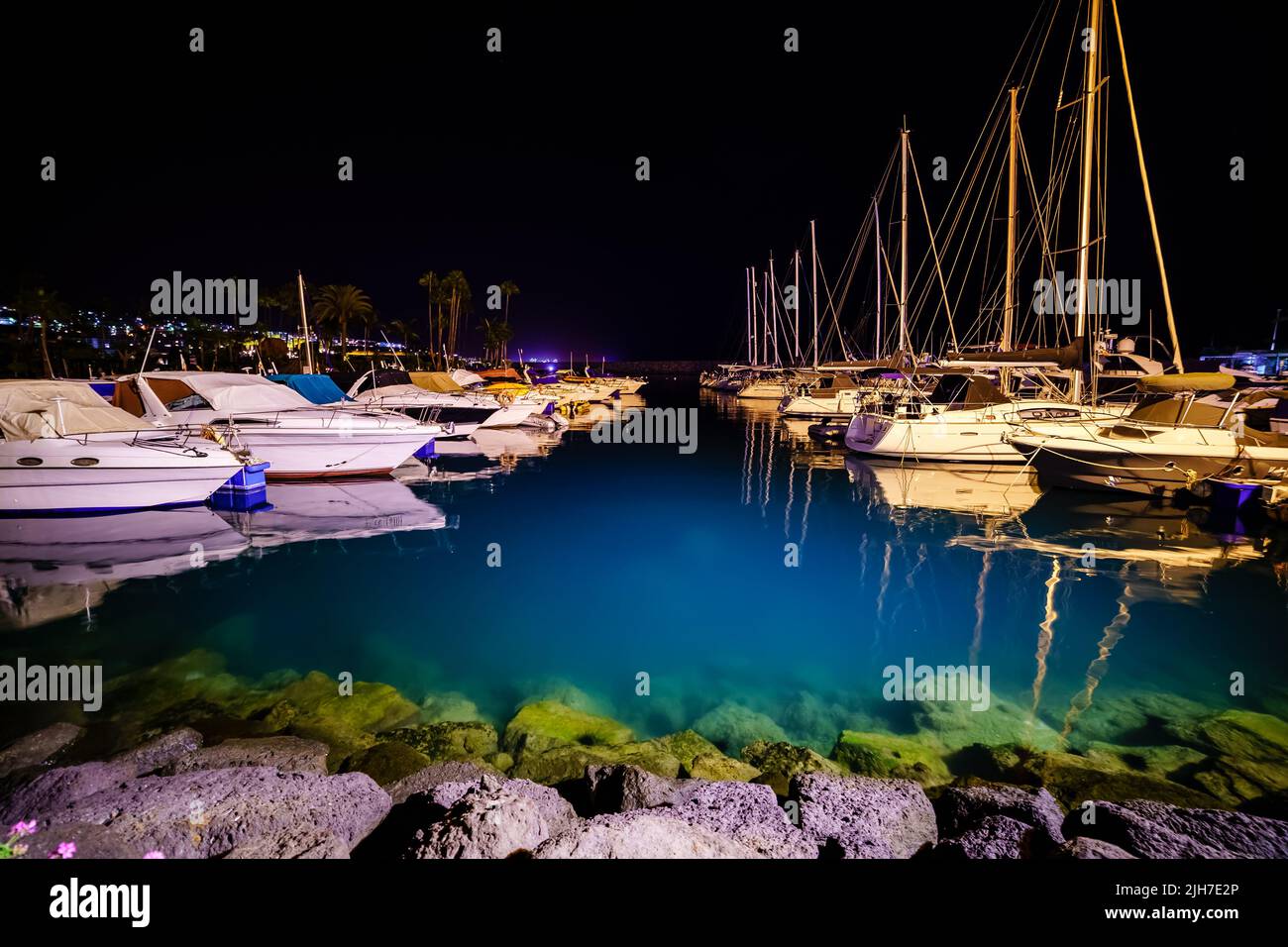 night photo of the marina with moored boats and transparent blue water that reveals the rocky bottom. Stock Photo