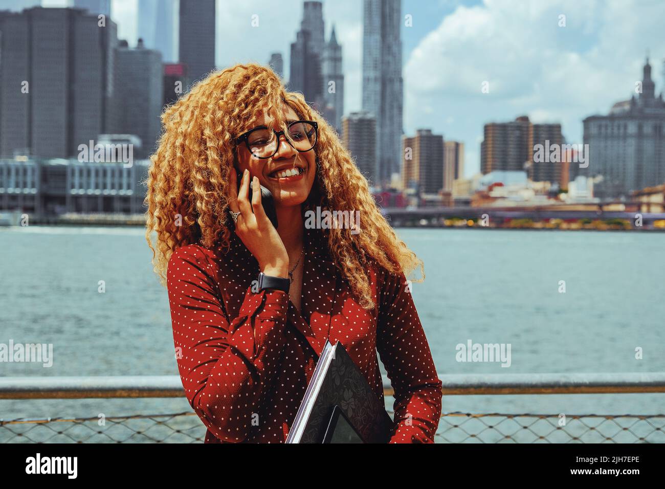 Portrait young adult entrepreneur millennial woman with eyeglasses and afro hair talking smiling on a phone call outdoors with Manhattan New York City skyline behind Hudson river Stock Photo