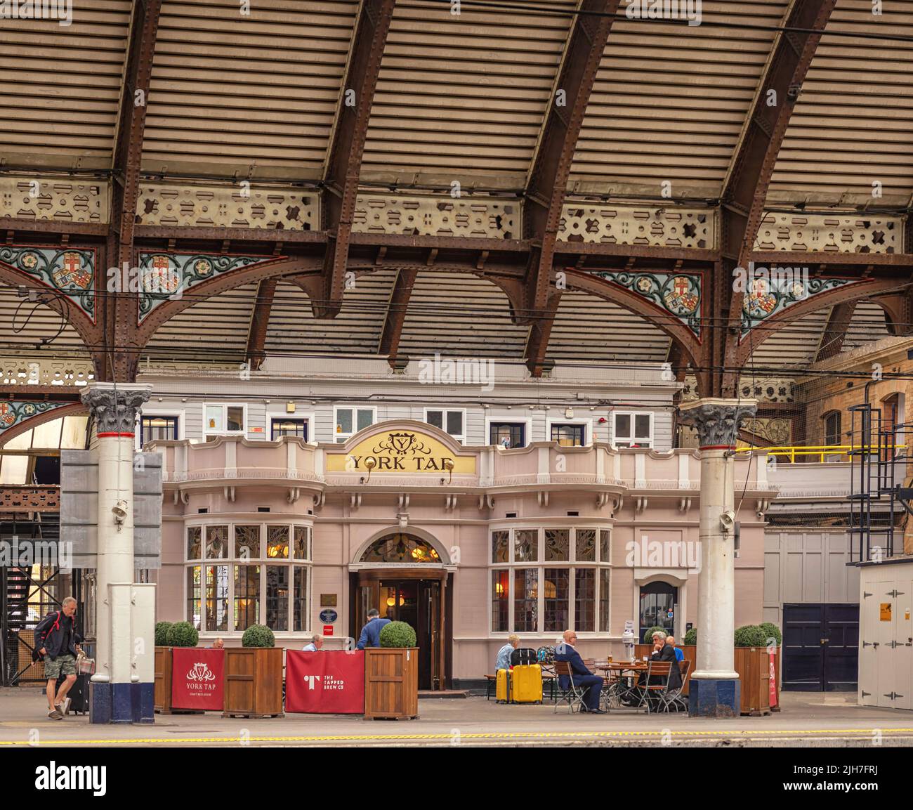 A pub is situated on a railway station platform under a historic canopy. People sit at tables and a passenger walks nearby. Stock Photo