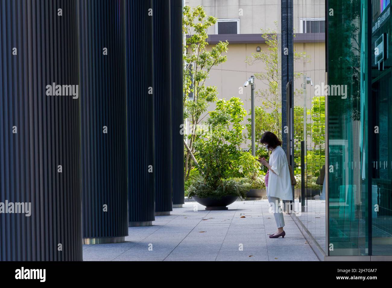 A Japanese woman uses a smartphone outside and modern office tower with pillars. Shibuya, Tokyo, Japan. Stock Photo