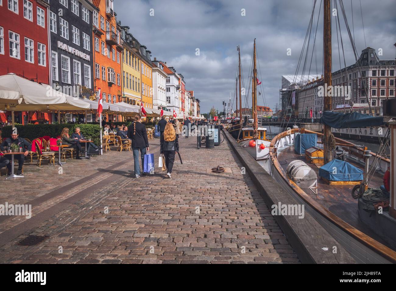 People walking and sitting on the terrace on Nyhavn street in Copenhagen along the bars, cafes and restaurants and wooden ships on the canal harbour. Stock Photo