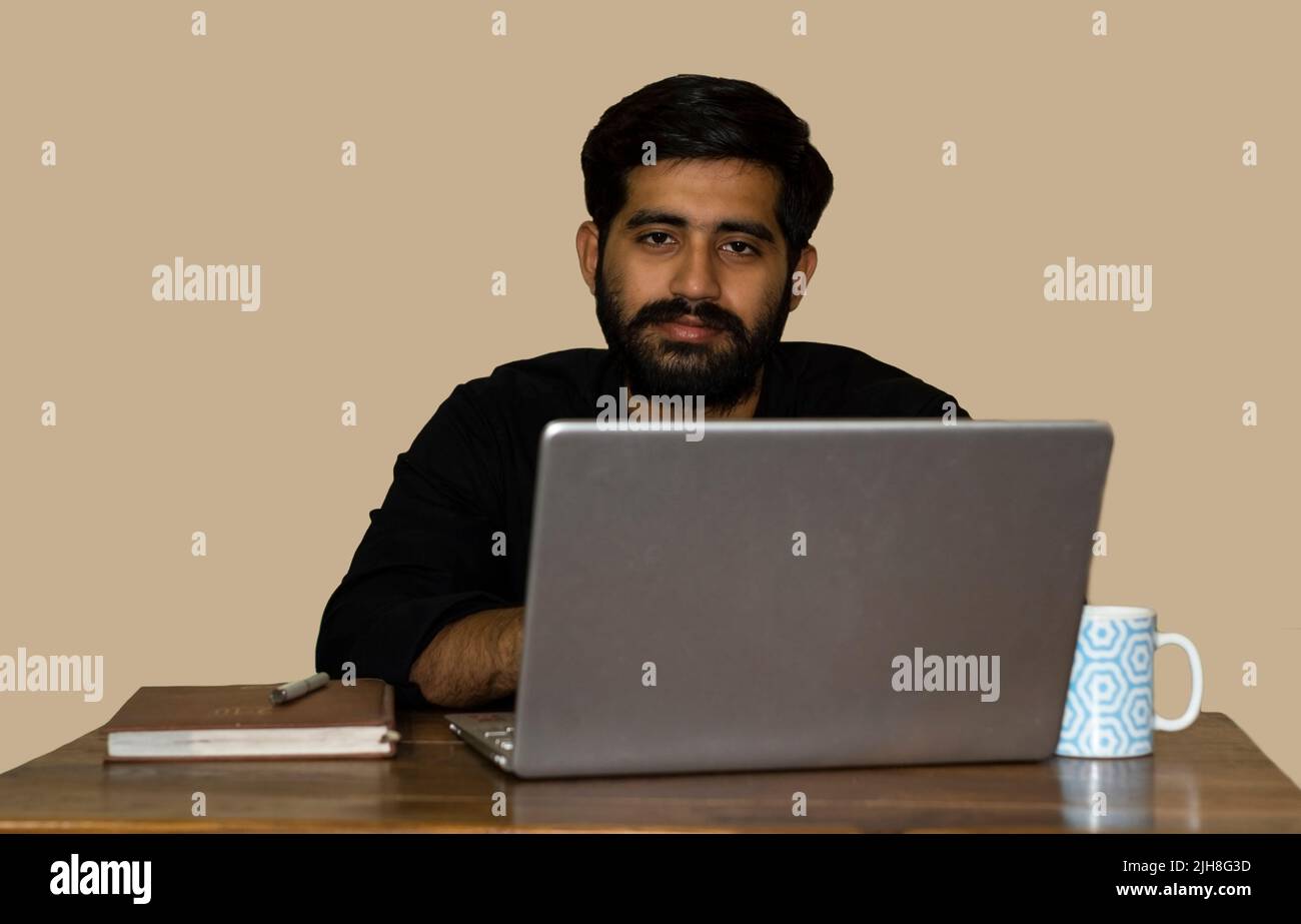 A young Indian man looking at camera while working on laptop. Selective focus on man's face. Stock Photo