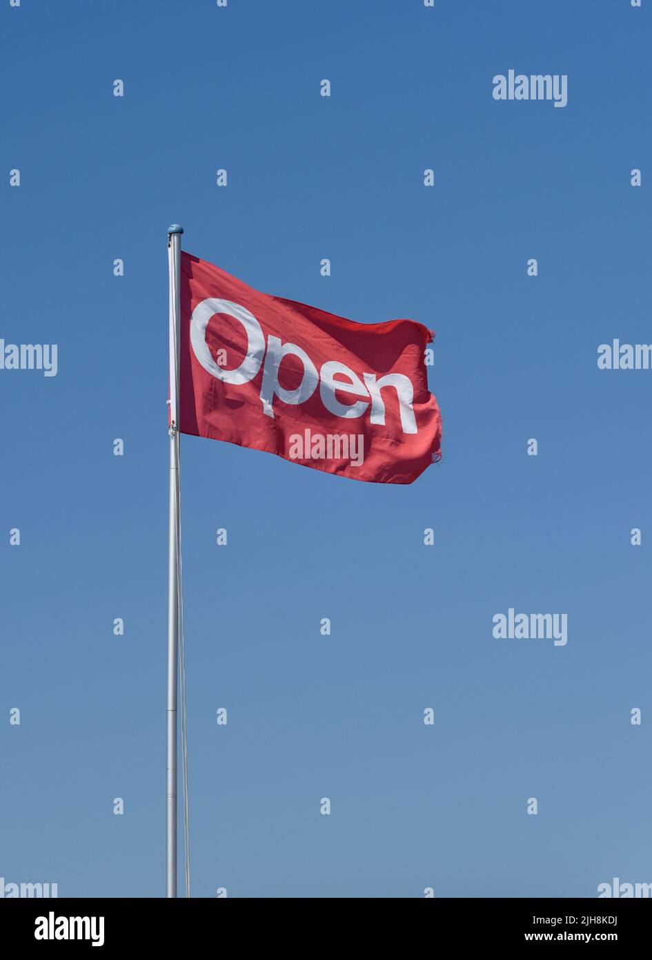 Open sign, red flag with white writing fluttering against a plain blue sky. Stock Photo