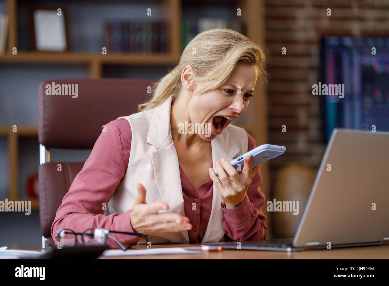 Furious businesswoman shouting expressively to smart phone while answering phone call at office. Angry annoyed woman enraged of bad service, having Stock Photo