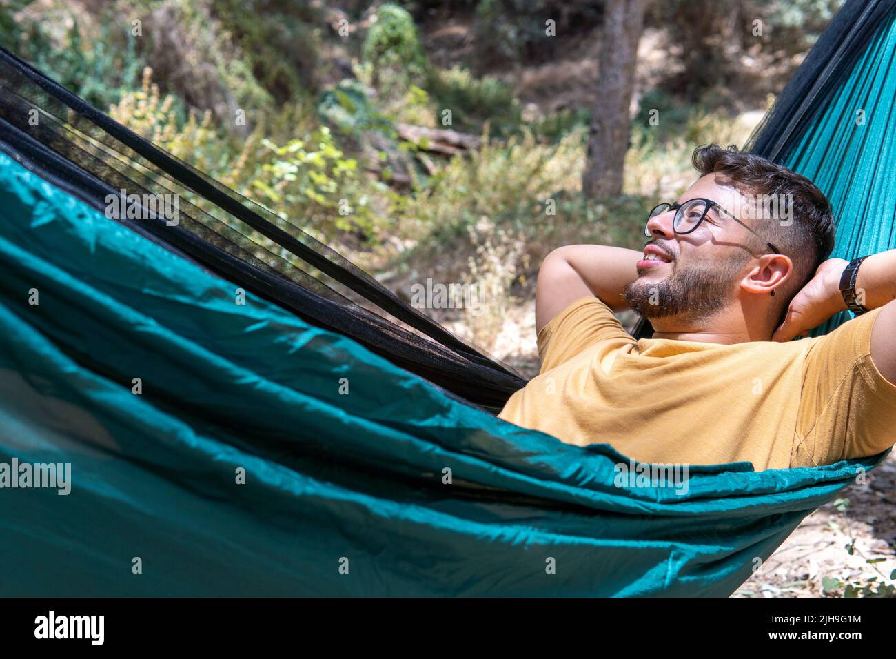 Close-up shot of a guy with glasses and yellow T-shirt lying in a green hammock enjoying nature in a forest Stock Photo