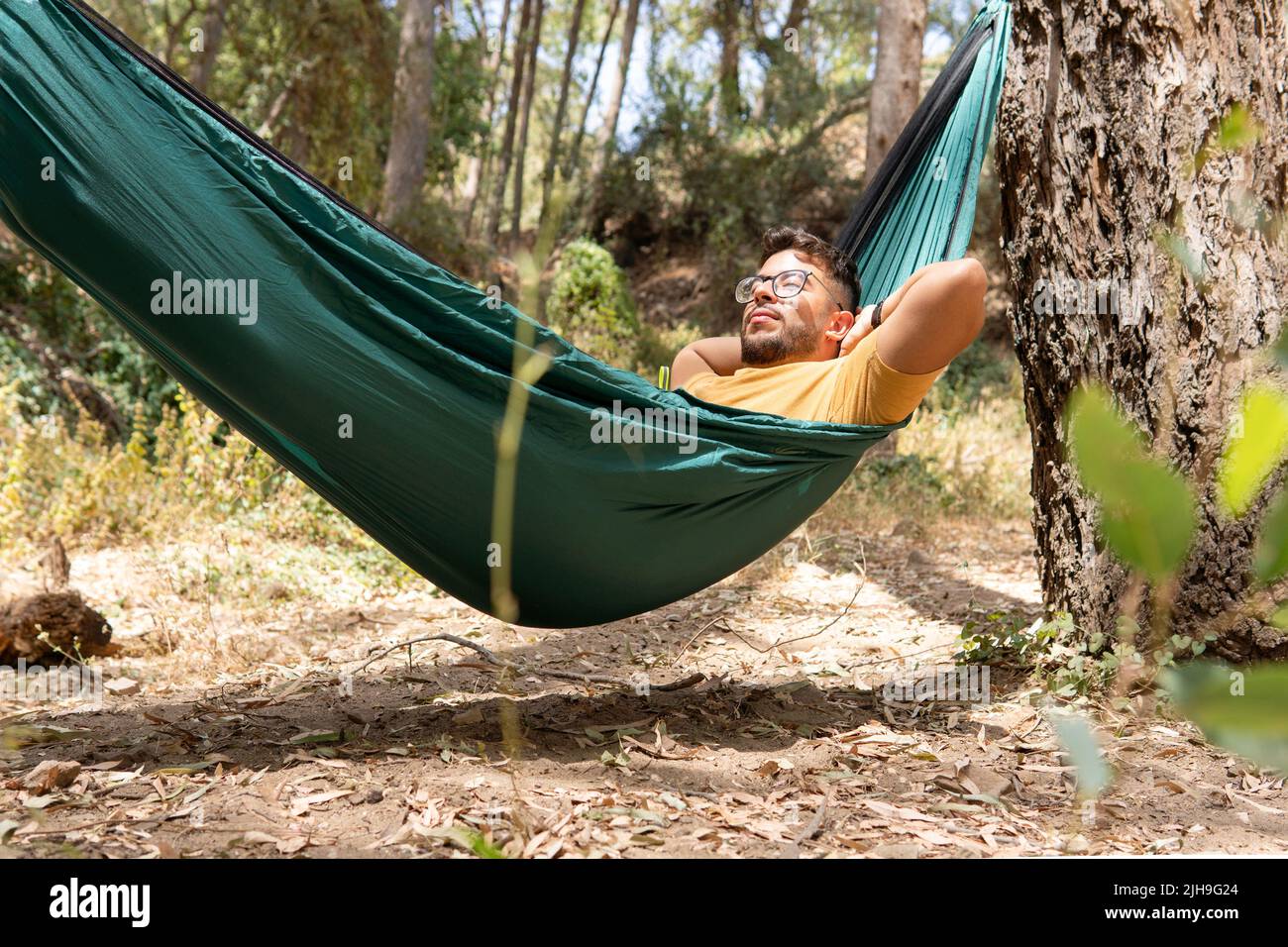 Open shot of a guy with glasses and yellow t-shirt lying in a green hammock enjoying nature in a forest Stock Photo