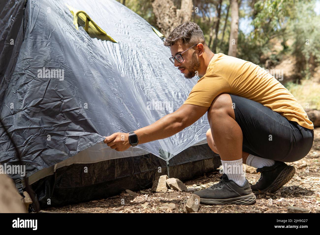 Young man in yellow T-shirt with glasses in a forest preparing his dark blue tent for camping in the forest. Stock Photo