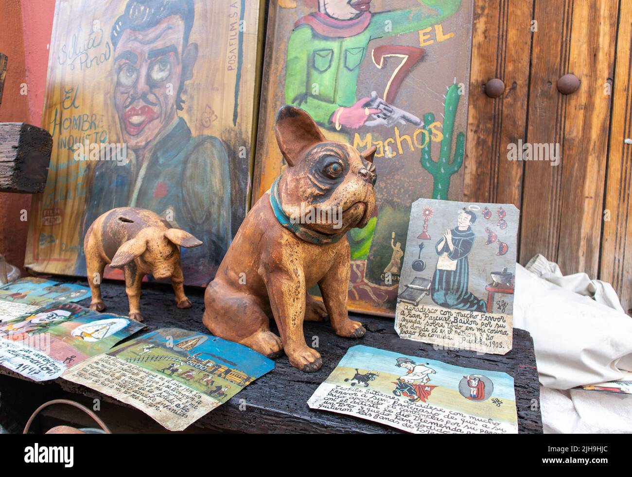 ceramic pottery dog at sale in a traditional mexican street market along with paintings catholic prayers with religious votive images and a piggy bank Stock Photo