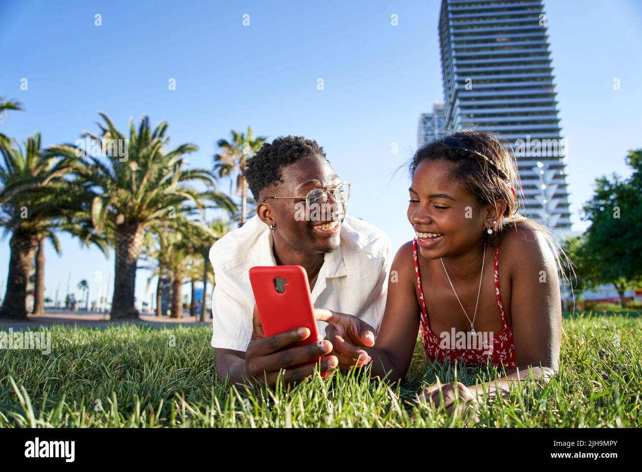A cheerful young African American couple using a mobile smart phone lying on the grass in the city park. Smiling boy and girl holding cell. Black Stock Photo