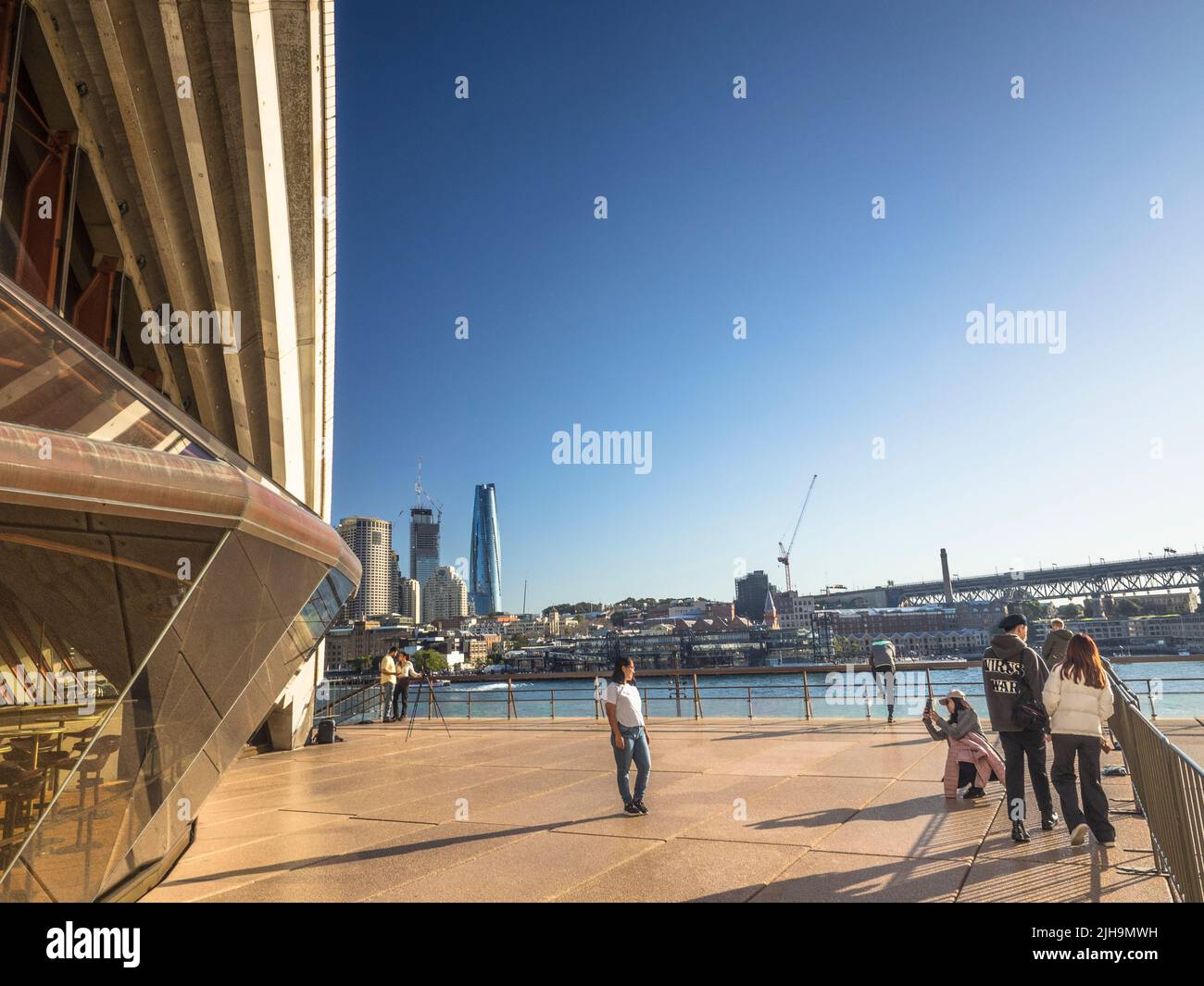 Tourists posing outside the restaurant shell of the Sydney Opera House. Circular Quay and the Sydney Harbour Bridge Southern Approach are in the bg. Stock Photo