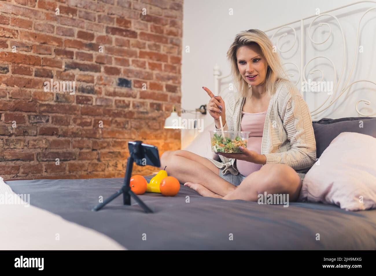young pregnant woman making a vlog and speaking about her vegetable salad, full shot bedroom healthy eating. High quality photo Stock Photo