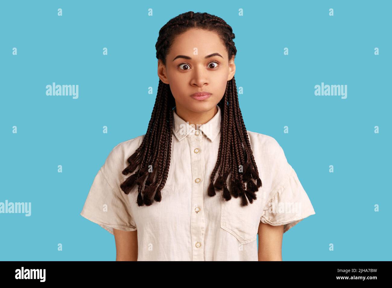 Portrait of funny silly woman with black dreadlocks looking at camera, cross eyed with stupid dumb face, has awkward confused comical expression. Indoor studio shot isolated on blue background. Stock Photo