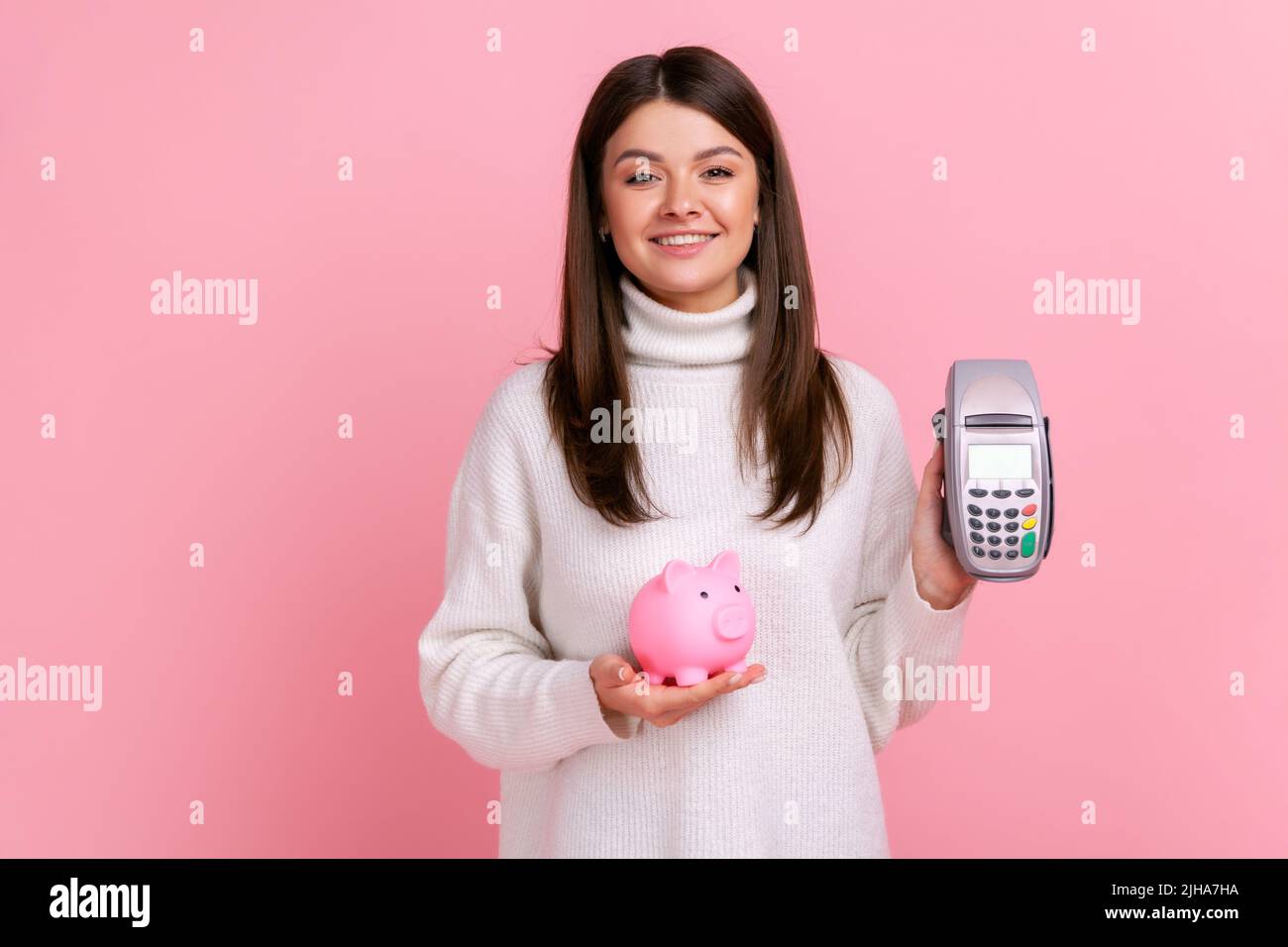 Pretty girl with dark hair showing pos payment terminal and piggybank, using cashless payments, nfc, wearing white casual style sweater. Indoor studio shot isolated on pink background. Stock Photo
