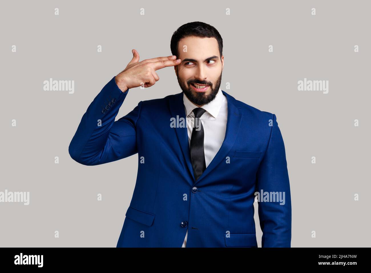 Portrait of bearded man holding fingers near head imitating gun, looking at camera with panic stressed expression, wearing official style suit. Indoor studio shot isolated on gray background. Stock Photo
