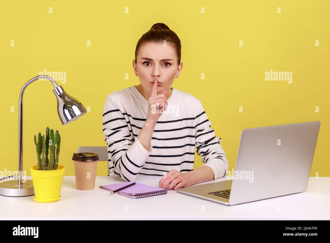 Be quiet. Serious woman in striped shirt sitting at workplace showing silence sign, looking at camera with finger on lips, asking to be silence. Indoor studio studio shot isolated on yellow background Stock Photo