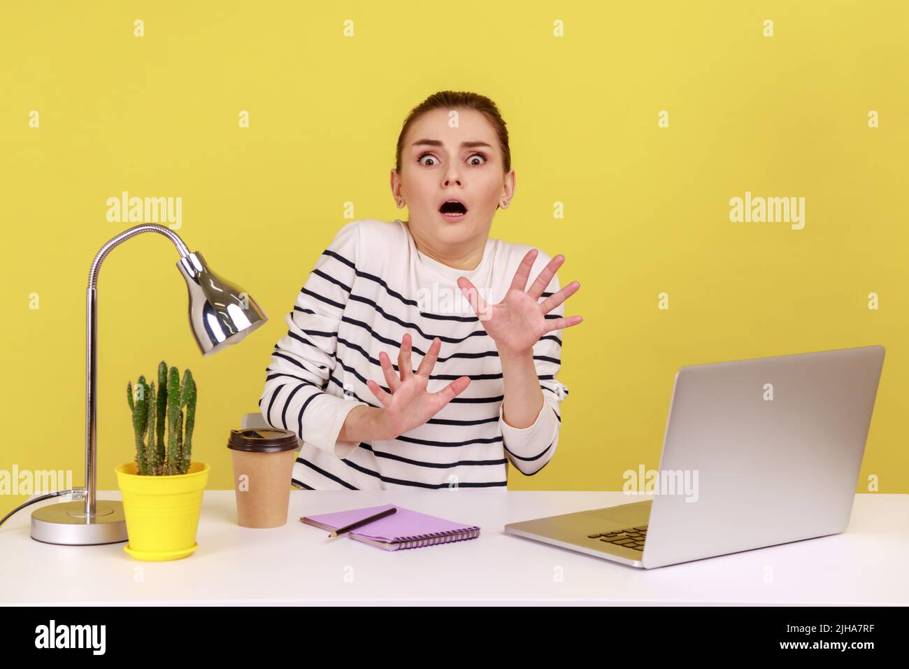 Frightened woman with shocked expression raising hands in stop gesture, defending herself, freaked out of troubles working on laptop. Indoor studio studio shot isolated on yellow background. Stock Photo