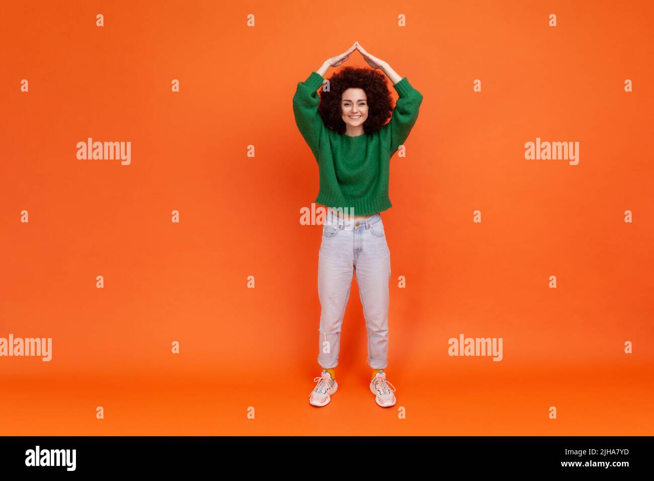 Full length portrait of woman with Afro hairstyle wearing green casual style sweater holding arms above head making roof gesture, insurance. Indoor studio shot isolated on orange background. Stock Photo