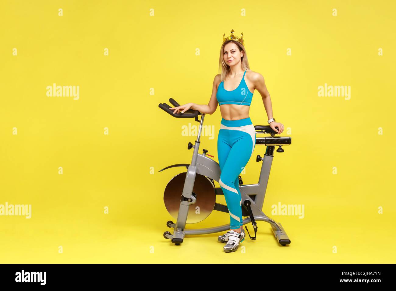 Full length portrait of confident slim woman standing near sport equipment with golden crown on her head, wearing blue sportswear. Indoor studio shot isolated on yellow background. Stock Photo