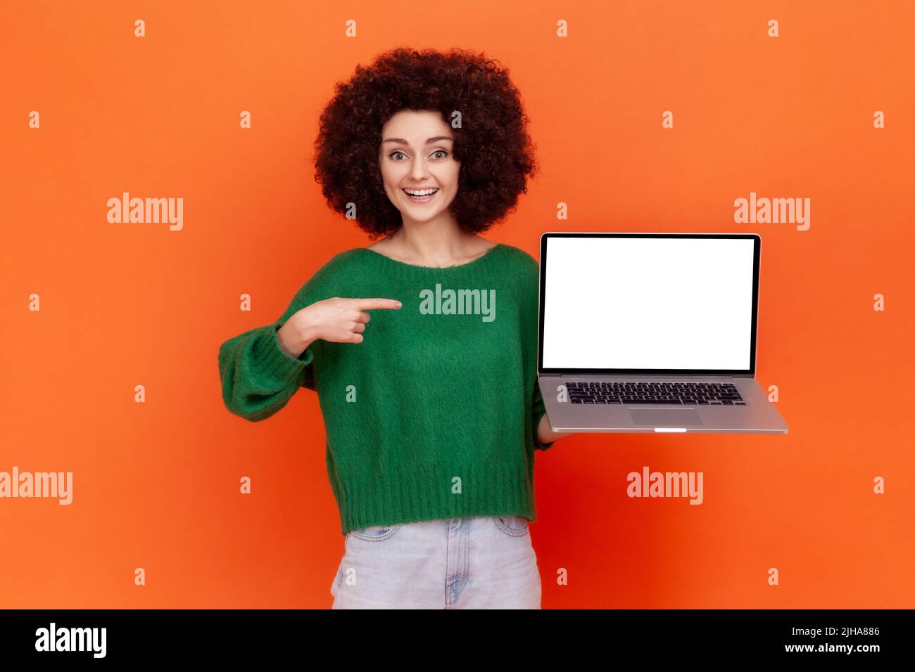 Good looking smiling woman with Afro hairstyle wearing green casual style sweater pointing at blank screen of laptop in her hands. Indoor studio shot isolated on orange background. Stock Photo
