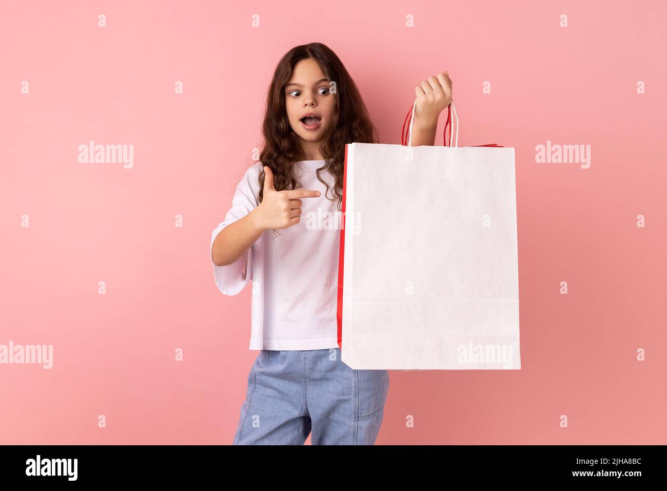 Portrait of shocked little girl wearing white T-shirt pointing at shopping bags, looking at camera with astonished facial expression. Indoor studio shot isolated on pink background. Stock Photo