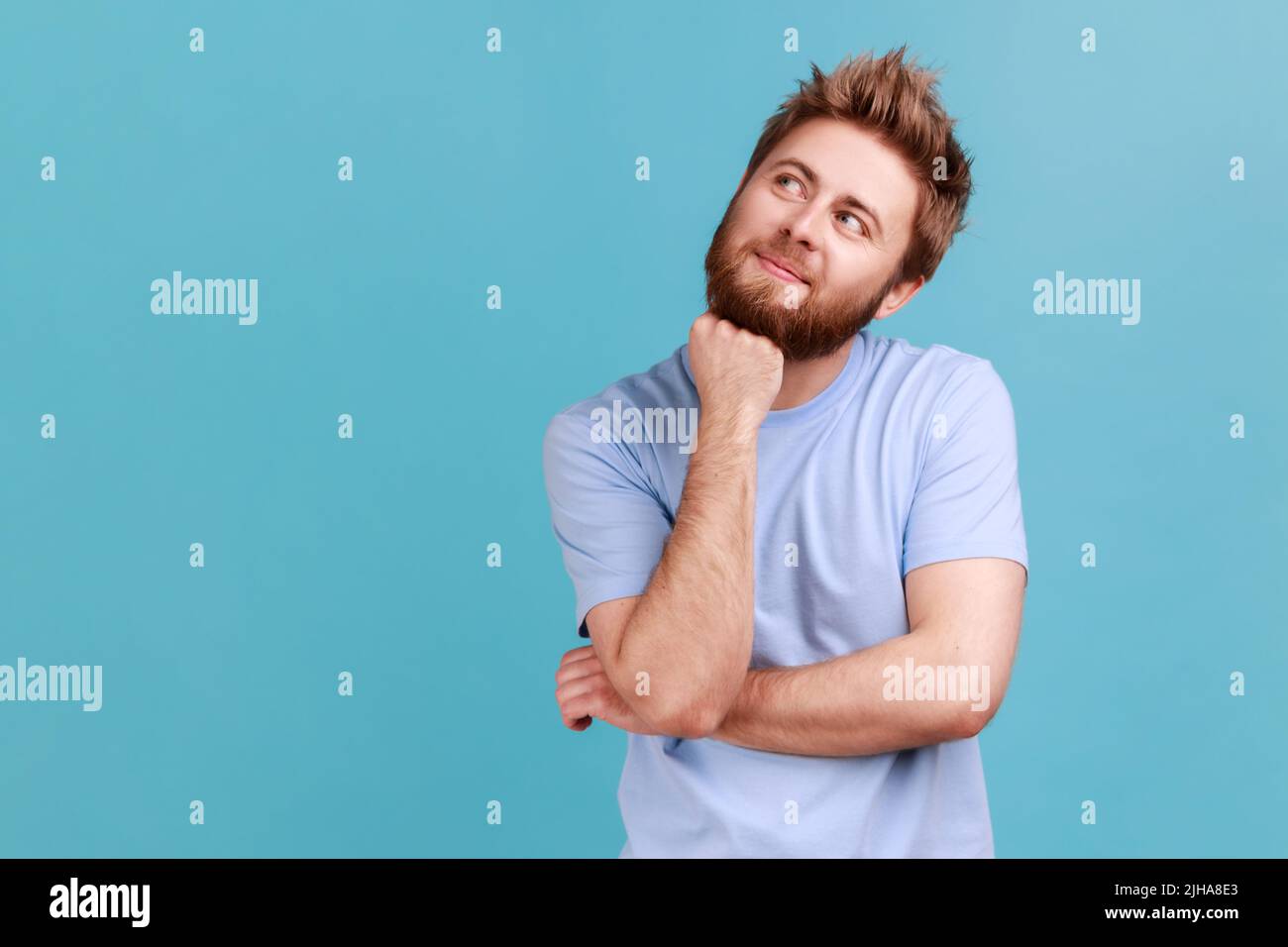 Portrait of smiling happy bearded man looking away with pensive expression, dreaming pleasant thoughts, fantasizing and making wish. Indoor studio shot isolated on blue background. Stock Photo