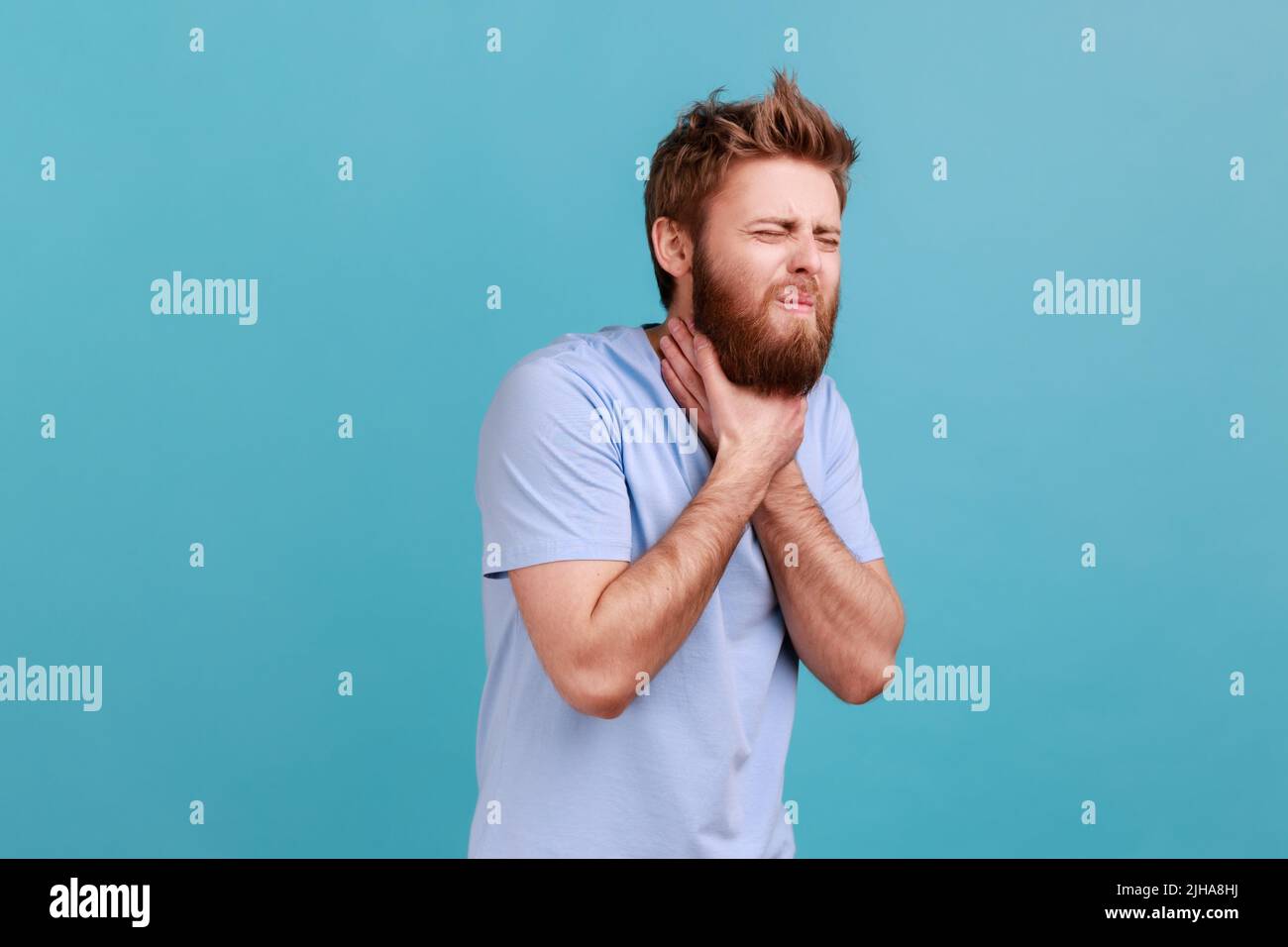 Portrait of bearded man grimacing touching his neck, feeling pain while swallowing, sore throat, risk of choking or anaphylactic shock. Indoor studio shot isolated on blue background. Stock Photo