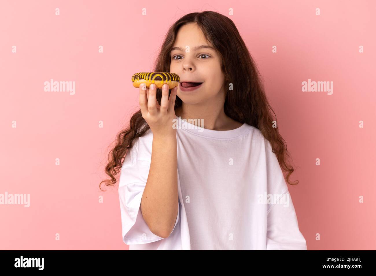 Portrait of delighted cute little girl wearing white T-shirt licking delicious donut, looking with desire to eat sweet dessert, showing tongue out. Indoor studio shot isolated on pink background. Stock Photo