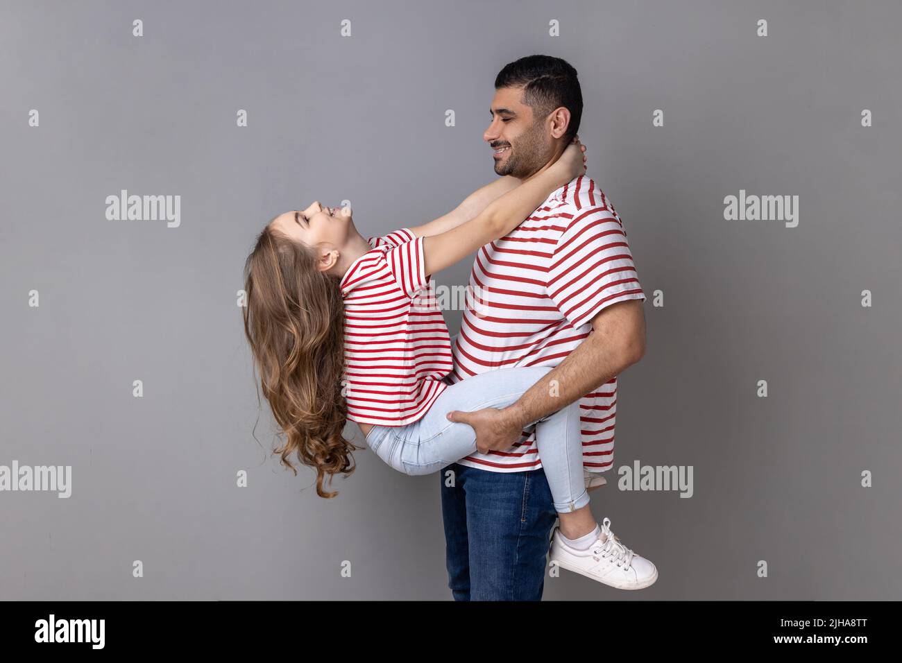 Portrait of delighted satisfied father and daughter in striped T-shirts spending time together, enjoying weekend, dad holding child. Indoor studio shot isolated on gray background. Stock Photo
