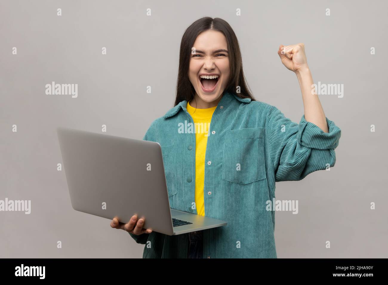 Extremely happy woman standing with laptop and clenching her fist, celebrating success, yelling happily, wearing casual style jacket. Indoor studio shot isolated on gray background. Stock Photo