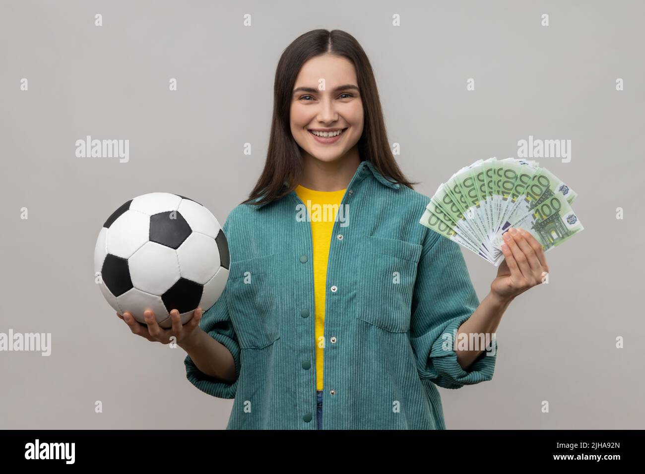 Portrait of satisfied woman showing soccer ball and fun of hundred euro bills, winning lot of money betting for sport, wearing casual style jacket. Indoor studio shot isolated on gray background. Stock Photo