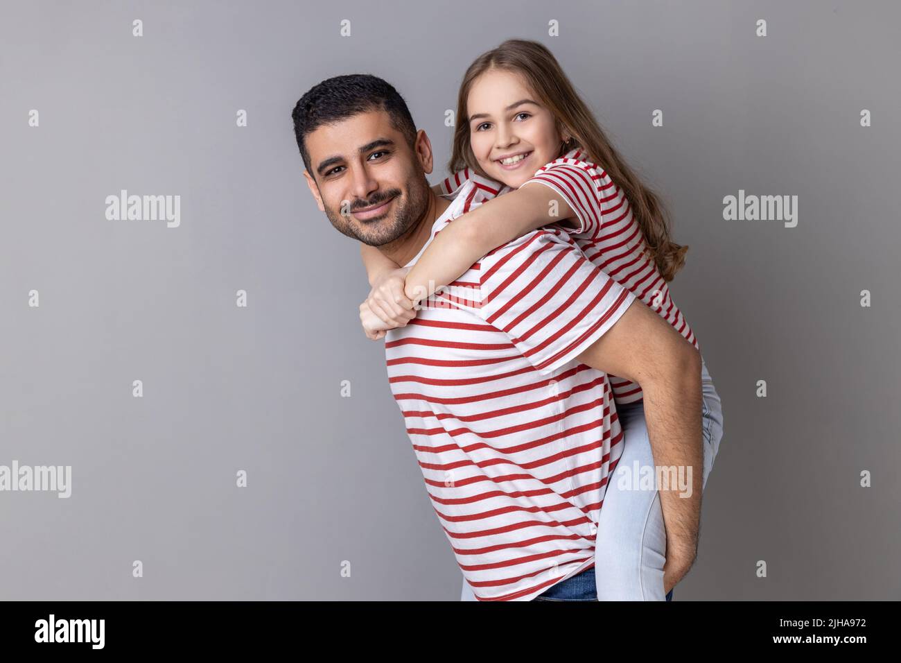 Portrait of smiling father and cute child in striped T-shirts posing together, dad give piggyback daughter, holding on back adorable kid. Indoor studio shot isolated on gray background. Stock Photo