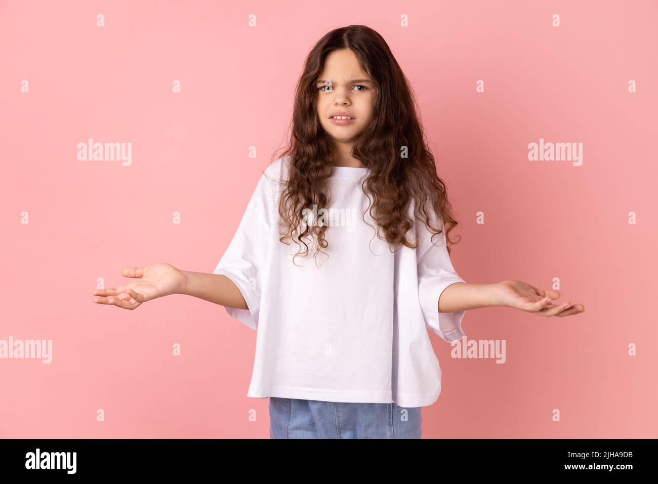 Portrait of annoyed frustrated little girl wearing white T-shirt standing with raised hands and indignant face asking why, annoyed by problem. Indoor studio shot isolated on pink background. Stock Photo
