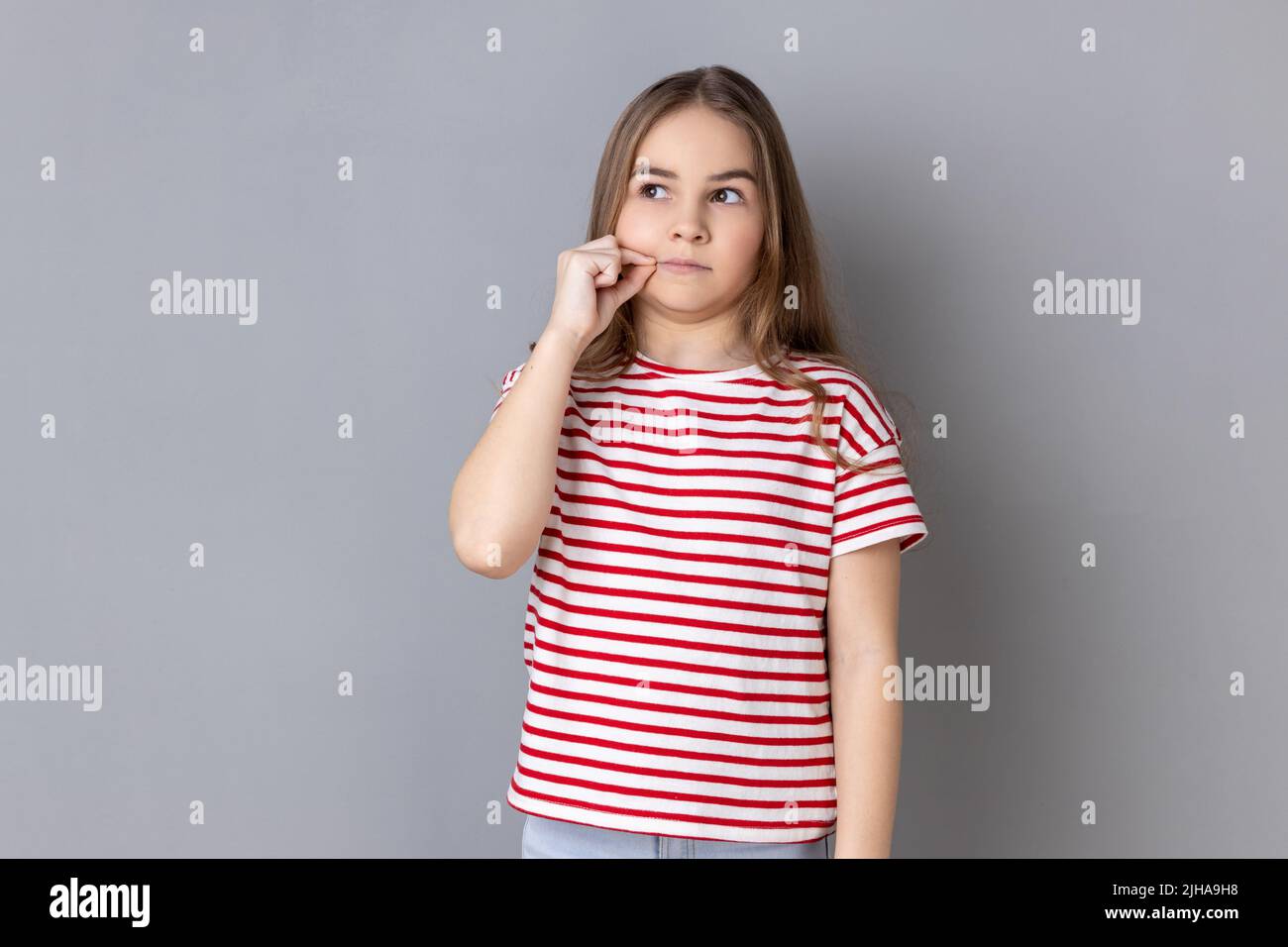 Portrait of adorable little girl wearing striped T-shirt having mystery look making zip gesture to close mouth, keeping secret, zipping lips. Indoor studio shot isolated on gray background. Stock Photo