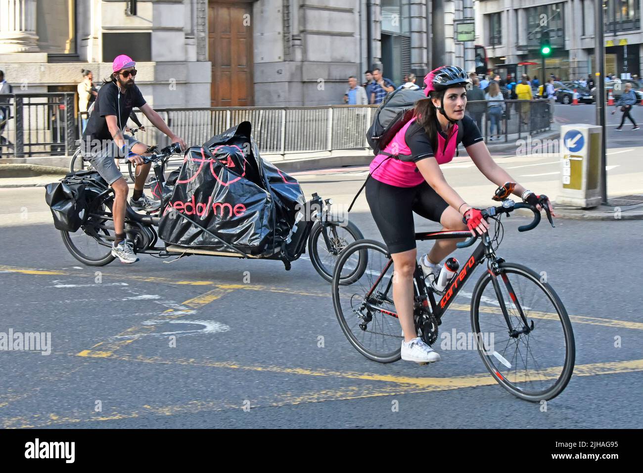 Woman cyclist with back pack riding drop handle bar bicycle across City of London box junction road followed by a man on long cargo bike England UK Stock Photo