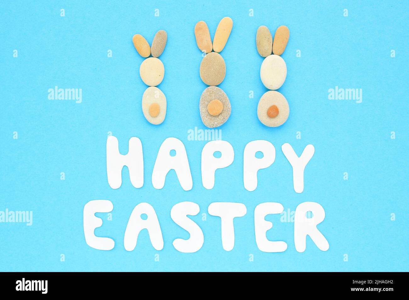 Happy Easter lettering and bunnies from pebbles on blue background. Flatlay postcard creative concept. Creative greetings with natural craft materials Stock Photo