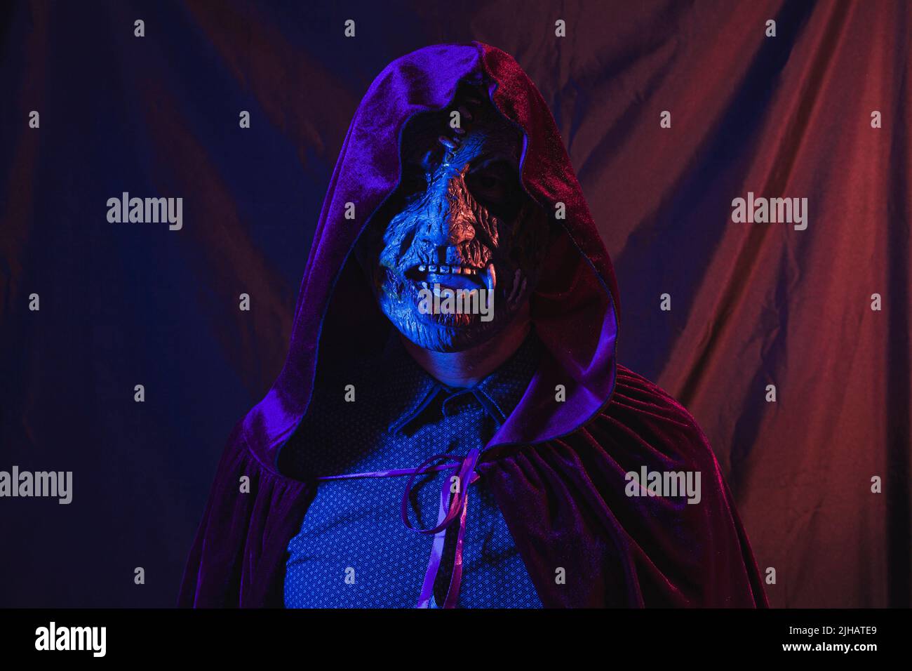 Portrait of a zombie dressed in a shirt and hooded cape facing the camera. The scene is dark, illuminated by blue and orange lights. Stock Photo