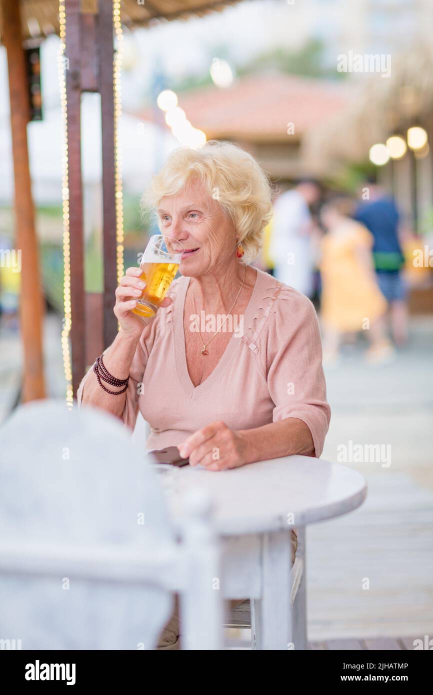 Mature woman sitting at a table in a summer cafe and drinking beer Stock Photo