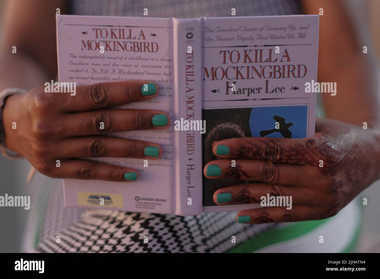 Black woman's hands with henna tattoo and aqua nails hold open the book, To Kill A Mockingbird, on legs wearing black and white houndstooth pants. Stock Photo
