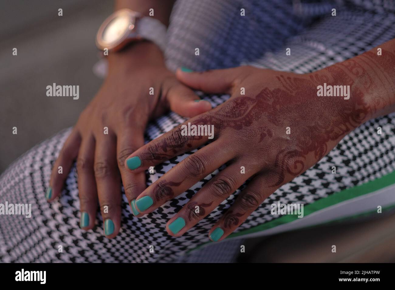 Black woman's hand with Arabic henna tattoo rests on crossed legs wearing black and white houndstooth pants with green stripe; nails are turquoise. Stock Photo