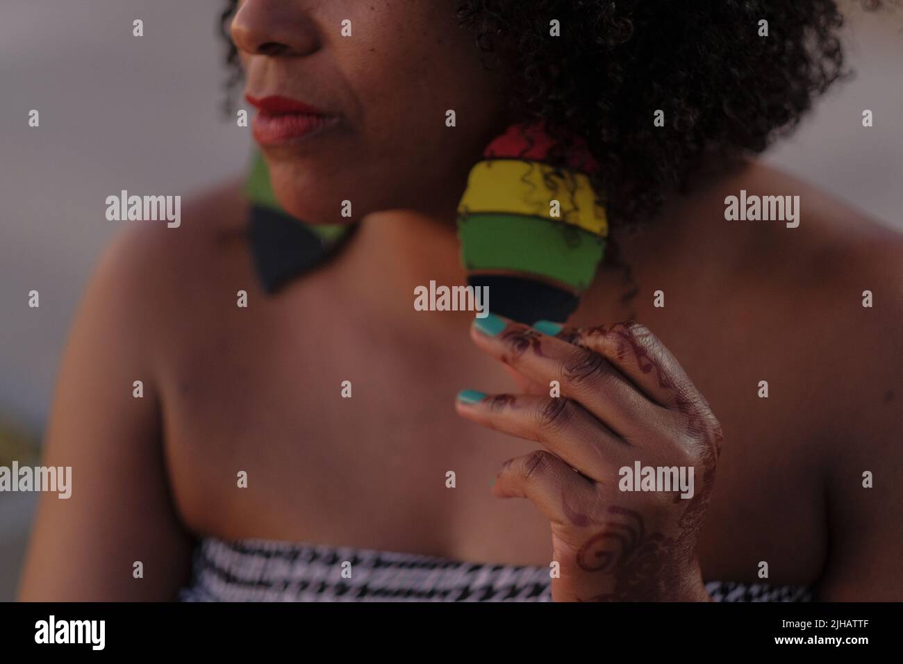 Black woman with afro hair holds big Rastafarian earring. Hand with Arabic henna tattoo and turquoise nails. Houndstooth tube top. Close crop of face. Stock Photo