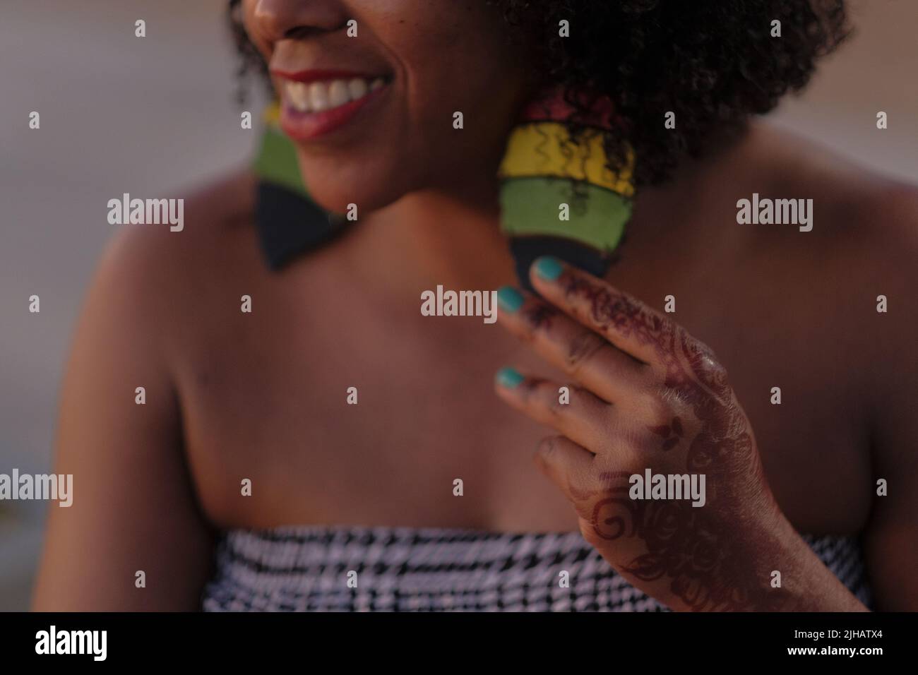 Smiling Black woman with afro hair holds big Rastafarian earring. Hand with henna tattoo and aqua nails. Houndstooth tube top. Close crop of face. Stock Photo