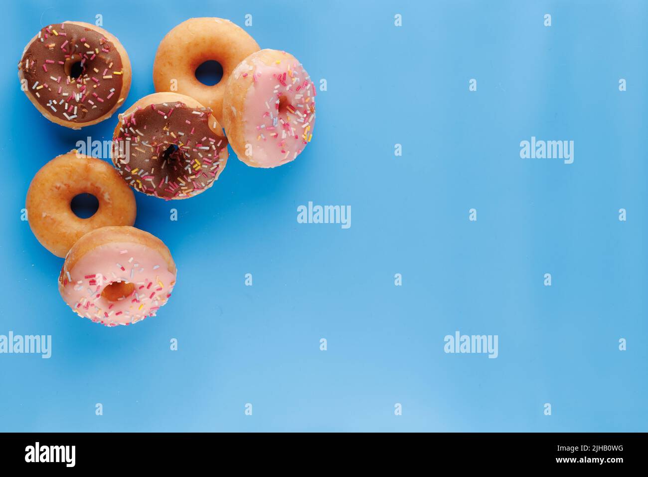 Sugar icing glazed doughnuts, including a chocolate sprinkle-topped variety, arranged in a flat lay on a solid background with negative space. Stock Photo