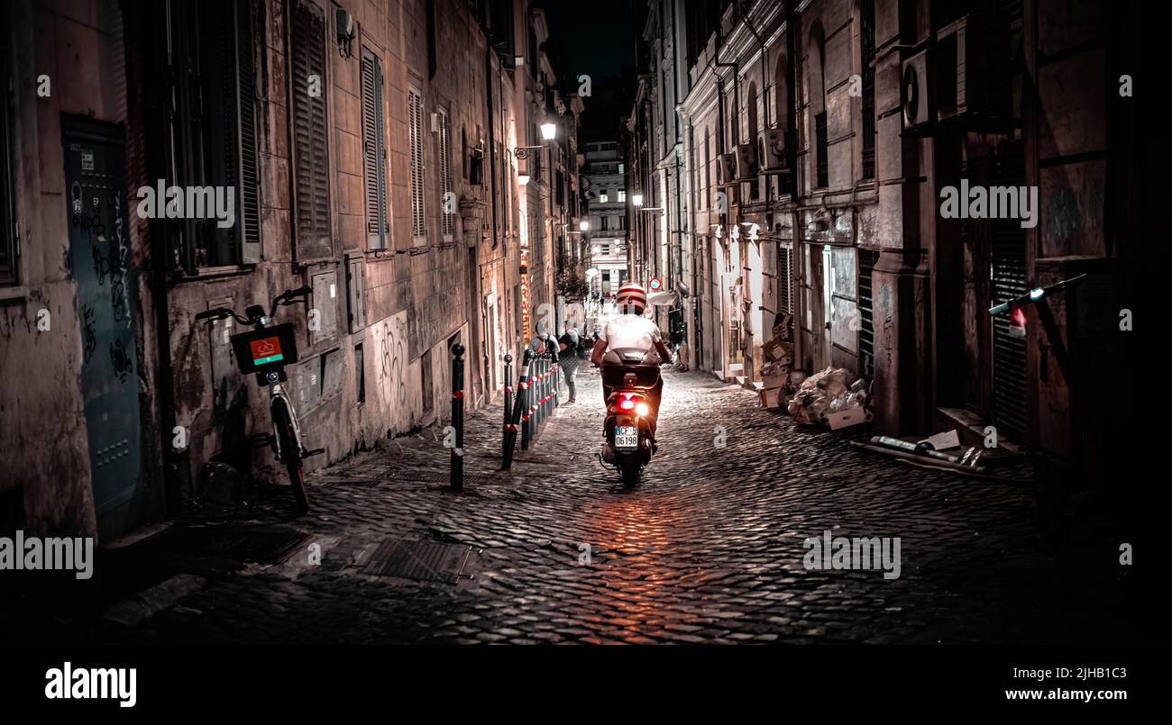 Rome, Italy - June 6, 2022: Person on motorbike rides away from camera down cobbled alley in Rome at night. Stock Photo