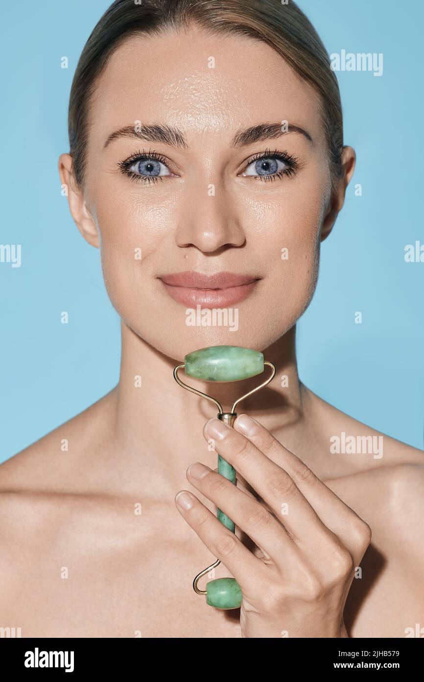 Young woman massages her face and chin using green jade roller for elastic perfect skin. facial jade roller, isolated on blue background Stock Photo