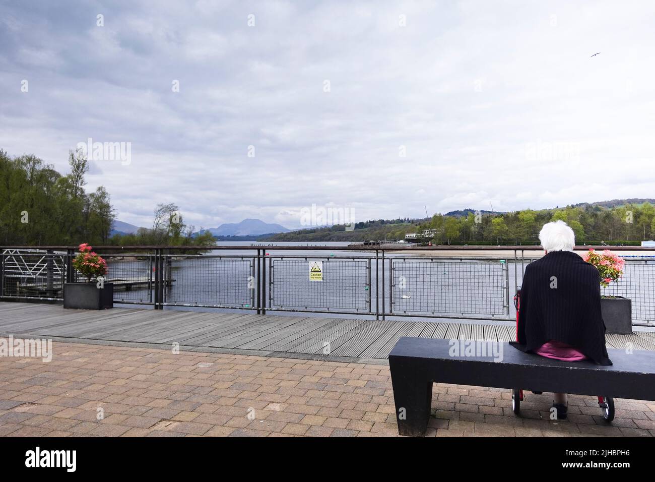 Elderly lady sits alone on bench looking out over Loch Lomond from Loch Lomond Shores Stock Photo